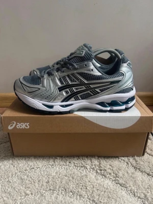 Asics skor  - Snygga Asics sneakers i silver och grå med meshpaneler och svarta detaljer. Skorna har en sportig design med vit och blå sula samt klassiska Asics-loggan på sidan. Perfekta för dig som gillar retrostil och komfort.