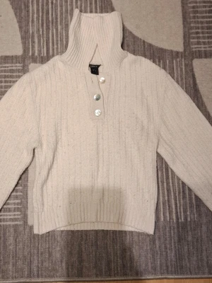 Beige stickad tröja från Lindex - Mysig beige stickad tröja från Lindex med hög krage och tre stora knappar framtill. Tröjan har ribbstickat mönster, långa ärmar och breda muddar. Perfekt för kalla dagar och enkel att styla till olika outfits.
