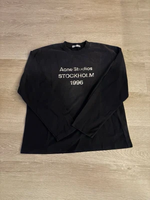 Svart långärmad tröja Acne Studios - Svart långärmad topp från Acne Studios med tryck i vitt framtill där det står 'Acne Studios STOCKHOLM 1996'. Klassisk rak passform och rund halsringning. Perfekt för dig som gillar stilrena och trendiga plagg.