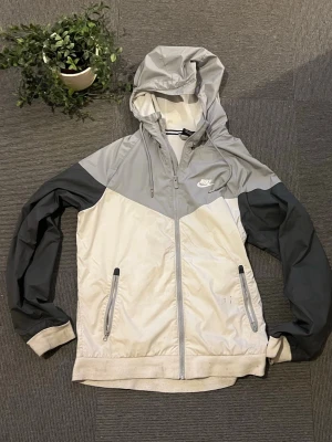 Nike vindjacka i grått och vitt - Snygg vindjacka från Nike i ljusgrått och vitt med mörkgrå ärmar. Jackan har huva, dragkedja framtill och två fickor med dragkedja. Tillverkad i lätt och vindtätt material, perfekt för blåsiga dagar. Nike-logga på bröstet för en sportig look.