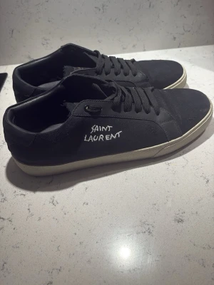 Svarta Saint Laurent sneakers - Svarta sneakers från Saint Laurent med vit broderad logga på sidan och vit sula. Skorna har klassisk låg profil, svarta skosnören och är tillverkade i canvas och läder. Perfekta för en clean och stilren look.