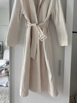 Ljusbeige trenchcoat från STOCKH LM - Snygg ljusbeige trenchcoat från STOCKH LM Studio i storlek XS. Klassisk modell med knytskärp i midjan och bred krage. Lång och rak passform, tillverkad i ett mjukt tyg med slätt foder. Perfekt för en stilren look. Som ny!! 