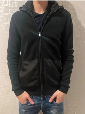 Svart cardigan - Svart cardigan från Roy robsson med hel dragkedja och två stora fickor framtill. Huvan är quiltad och ger en cool kontrast till resten av tröjan. Perfekt för dig som gillar en enkel men snygg stil.