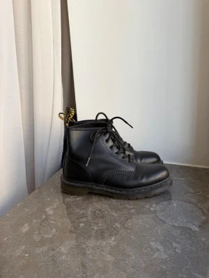 Dr. Martens 101 kängor strl. 41 - Säljer ett par svarta kängor i läder med snörning och grov sula. Modellen har en ikonisk gul flärp bak och rundad tå. Perfekta för dig som gillar en tidlös och edgy stil. Passar till många olika outfits och är riktigt bekväma.