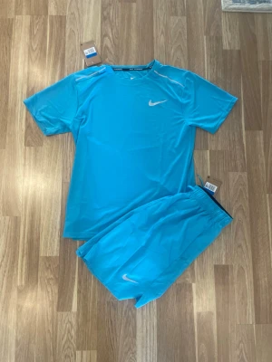 Blått träningsset från Nike - Snyggt blått träningsset från Nike med matchande t-shirt och shorts. Båda plaggen har Nike-loggan i vitt och är gjorda i ett lätt och snabbtorkande material, perfekt för träning eller sportaktiviteter. T-shirten har rund hals och shortsen har resår i midjan.