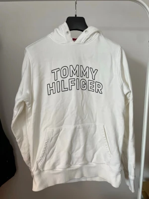 Vit Tommy Hilfiger hoodie - Snygg vit hoodie från Tommy Hilfiger med stor logga framtill. Klassisk känguruficka och ribbade muddar. Tillverkad i mjukt material och har huva med dragsko. Perfekt för en avslappnad och trendig look.