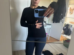 Svart långärmad tröja med knappar - Säljer en svart långärmad tröja med tight passform och dekorativa silverfärgade knappar vid ärmsluten. Tröjan har rund halsringning och är gjord i ett mjukt, stretchigt material som sitter snyggt på kroppen.