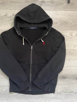 Polo Ralph Lauren Zip Hoodie – Storlek M - Skick: Bra begagnat skick.  Passform: Medium (M).  Material: Mjuk bomullsblandning med skön struktur i luvan.  Detaljer: Svart huvtröja med hel dragkedja, röd klassisk broderad Polo-logotyp på bröstet, vita dragskor samt delad känguruficka framtill. Skickas snabbt och spårbart 