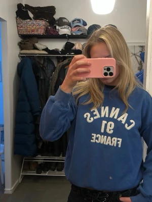Blå sweatshirt med tryck - Blå sweatshirt med vit text 'CANNES 91 FRANCE' på bröstet. Tröjan har rund halsringning och långa ärmar. Perfekt för en avslappnad och cool stil.