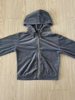 Grå velourhoodie med dragkedja - Mjuk och skön hoodie i grå velour med dragkedja framtill och justerbar huva med snören. Klassisk passform och ribbade muddar vid ärmslut och nederkant. Perfekt för en avslappnad och trendig look.