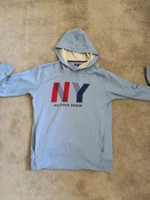 Ljusblå Hilfiger Denim hoodie - Snygg ljusblå hoodie från Hilfiger Denim med stort NY-tryck i rött och blått på bröstet. Hoodien har en mjuk, ljusgrå insida i huvan och små Hilfiger-detaljer. Tillverkad i bomullsmix och har ribbade muddar vid ärmslut och nederkant.