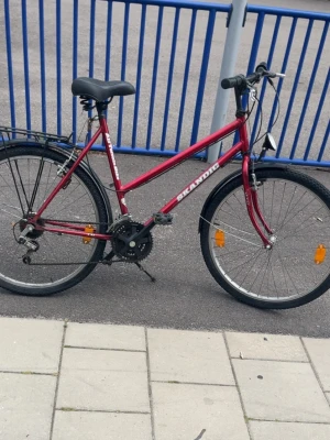 Röd cykel från Skandic - Säljer en röd cykel från Skandic med svart sadel, pakethållare och reflexer på hjulen. Cykeln har växlar, handbromsar och en klassisk ram i metall. Perfekt för dig som vill ta dig runt smidigt i stan. Back däcken behöver bytas bara eller pumas