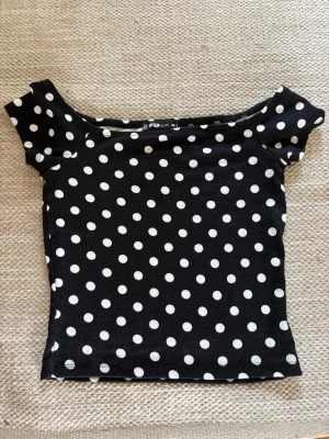 Svart prickig topp med bred urringning - Säljer en svart topp med vita prickar. Toppen har korta ärmar och är figurnära i modellen. Perfekt för dig som gillar retrostil och vill sticka ut lite extra. Materialet känns mjukt och stretchigt.