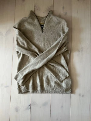 Stickad tröja med halvzip beige  - Mysig beige stickad tröja med halvzip och krage. Tröjan har långa ärmar och ribbade muddar vid ärmslut och nederkant. Perfekt för lager på lager och enkel att matcha med olika outfits.