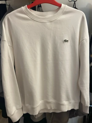 Vit sweatshirt från Lacoste - Klassisk vit sweatshirt från Lacoste med rund halsringning och det ikoniska krokodilmärket broderat på bröstet. Tröjan har ribbade muddar vid ärmslut och nederkant. Perfekt för en clean och stilren look.