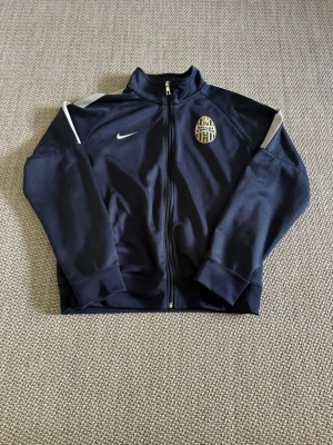 Mörkblå Hellas Verona Nike träningsjacka - Mörkblå träningsjacka från Nike med Hellas Verona-logga på bröstet. Jackan har hel dragkedja, ståkrage och vita detaljer på axlarna. Perfekt för fotbollsträning eller som sportig vardagsstil.