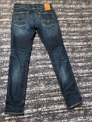 Mörkblå hyperflex Replay jeans, slim fit - Säljer ett par mörkblå hyperflex slim fit jeans från Replay med snygga slitningar och klassiska bakfickor med vågdetalj. Jeansen har normal midja och är tillverkade i bomull med lite stretch för extra komfort. Priset kan diskuteras. Skriv om ni var några funderingar!