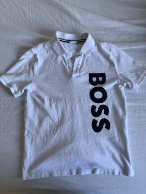 Vit BOSS t-shirt med tryck - Säljer en vit t-shirt med klassisk krage och svart BOSS-tryck längs sidan framtill. T-shirten har korta ärmar och är gjord i mjuk bomull som känns skön mot huden. Perfekt för en clean och stilren look. Pris går att diskutera 