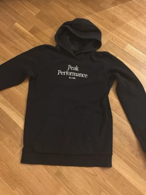 Svart Peak Performance hoodie - Svart hoodie från Peak Performance med vit logga på bröstet. Klassisk modell med huva och stor magficka framtill. Mjuk bomullskänsla och ribbade muddar vid ärmslut och nederkant. Perfekt för en chill och sportig stil.