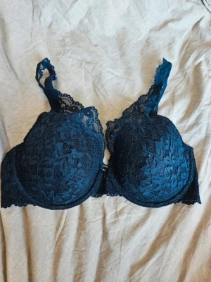 Mörkblå spets-bh med vaddering - Snygg mörkblå bh med vadderade kupor och bygel. Bh:n är täckt av blommig spets och har justerbara axelband samt hakknäppning bak. Perfekt för dig som gillar en feminin och bekväm stil.