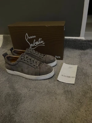 Gråa sneakers från Christian Louboutin - Snygga gråa sneakers från Christian Louboutin i mocka med klassisk låg profil och vita sulor. Skorna har matchande grå skosnören och diskreta sömmar för en clean look. Perfekta för dig som vill ha något stilrent och exklusivt.