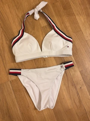 Vit bikini från Tommy Hilfiger - Vit bikini från Tommy Hilfiger med sportig känsla. Både överdel och underdel har breda band med röd, vit och marinblå randig detalj. Överdelen knyts i nacken och har vadderade kupor. Klassisk och stilren design, perfekt för stranden.