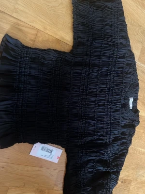 Svart frillad polokrageblus - Trendig svart blus med frillad struktur och polokrage. Blusen har långa ärmar och är tillverkad i ett glansigt, stretchigt material som ger en snygg passform. Perfekt för dig som gillar en unik och modern look.