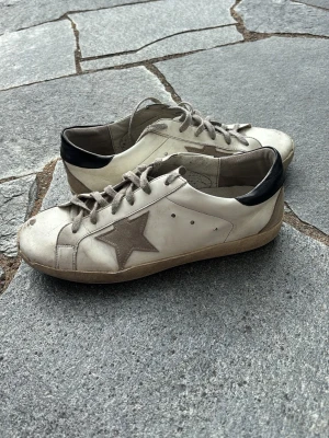 Golden goose - Snygga vita sneakers med beige stjärna på sidan och svart hälparti. Skorna har beige snörning och en klassisk låg siluett. Materialet är läder med mockadetaljer och sulan är i gummi. Perfekta för en avslappnad streetstyle-look.