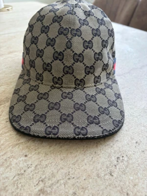 Gucci beige keps med GG-mönster - Säljer en beige keps från Gucci med det klassiska GG-monogrammet i svart över hela kepsen. Kepsen har böjd skärm och är tillverkad i ett slitstarkt tygmaterial. Perfekt för dig som vill ha en stilren och lyxig accessoar.