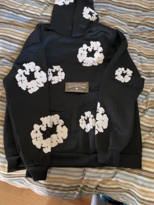 denim tears hoodie - Säljer en svart hoodie med stora vita blommönster tryckta över hela plagget, inklusive huvan. Hoodien har en klassisk känguruficka framtill och ribbade muddar vid ärmar och nederkant. Perfekt för dig som vill sticka ut med en unik och trendig stil.