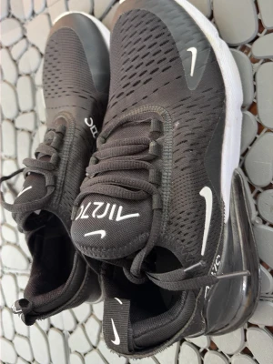Nike Air Max 270 svarta sneakers - Svarta Nike Air Max 270 sneakers med vit sula och tydlig Air Max-bubbla i hälen. Ovandelen är i meshmaterial för bra ventilation och har vita detaljer samt logga på sidan och plösen. Yttersulan har röd detalj undertill. Snygg och sportig design.