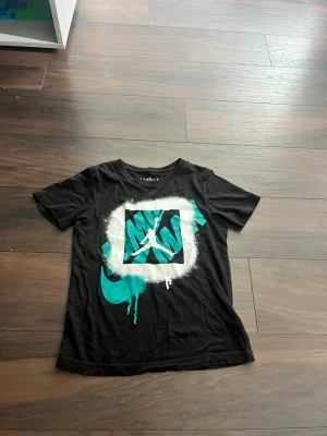 Jordan svart t-shirt med grafiskt tryck - Säljer en svart t-shirt från Jordan med ett stort grafiskt tryck framtill i turkos, vitt och svart. Trycket har en cool spray paint-effekt och visar den klassiska Jumpman-loggan. T-shirten har rund hals och korta ärmar, perfekt för en streetwear-look.
