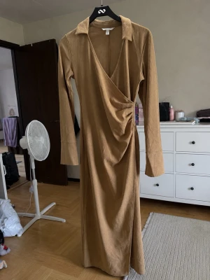 Lång brun omlottklänning - Snygg långärmad omlottklänning i brun färg med krage och djup v-ringning. Klänningen har en draperad detalj vid midjan och är i ett mjukt material som ger en elegant look.