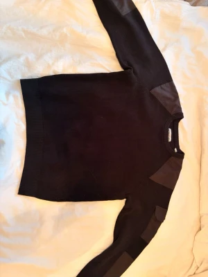 Svart stickad tröja med detaljer - Svart stickad tröja med förstärkta partier i tyg på axlar och armbågar. Tröjan har rund halsringning och långa ärmar. Materialet ser ut att vara en tjockare stickning, perfekt för kyligare dagar. Enkel och stilren design som passar till det mesta.