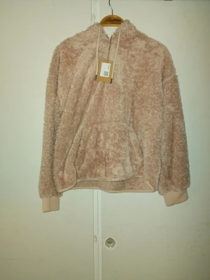 Beige fluffig hoodie med huva - Mysig beige hoodie i fluffigt teddy-material med huva och dragsko. Långa ärmar med ribbade muddar och en stor magficka framtill. Perfekt för dig som gillar en avslappnad och trendig stil. Storlek S