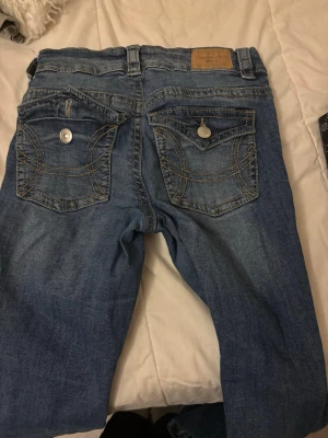 Blå jeans med ficklock och kontrastsömmar - Säljer ett par blå jeans med snygga kontrastsömmar från Gina, jeansen har två bakfickor med lock och silverfärgade knappar. Klassisk denimlook med slitningar och normalhög midja. Perfekta till en avslappnad stil.