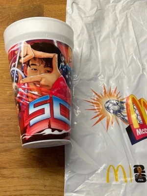 McDonald's FIFA plastmugg - Stor plastmugg från McDonald's med färgglad FIFA-design. Motivet visar en person i röd jacka, sporttema och stora bokstäver. Muggen har vit kant och lock. Perfekt för samlare eller sportfans.