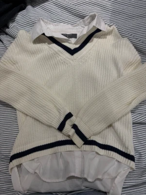 Vit stickad tröja med skjortkrage - Snygg vit stickad tröja från Primark med klassisk v-ringning och mörkblå ränder vid hals, ärmslut och nederkant. Tröjan har en fastsydd vit skjortkrage och skjortkant nedtill för en preppy look. Perfekt till jeans eller kjol.