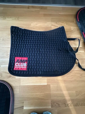 Svart schabrak Jump Club - Säljer ett svart, quiltat schabrak från Jump Club med röd och vit logga på sidan. Perfekt till ridning och har praktiska fästen för sadel och gjord. Materialet är slitstark bomull och designen är klassisk och stilren till ponny💗hämtas här Malmö!💗
