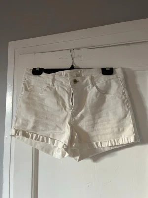 Vita denimshorts med fickor - Snygga vita shorts i denim med klassisk femficksmodell, dragkedja och knapp framtill. Shortsen har bälteshällor och uppvikta benslut för en avslappnad look. Perfekta till sommaren och lätta att matcha med olika stilar.