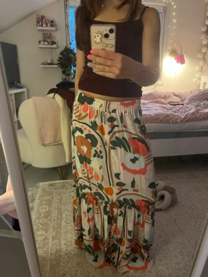 Färgglad blommig långkjol - Säljer en långkjol med stort blommigt mönster i grönt, orange, rött och vitt. Kjolen har resår i midjan och volang nertill för en flowy känsla. Perfekt för dig som gillar färg och vill sticka ut med din stil. Stretchig midja så går till både xs-m