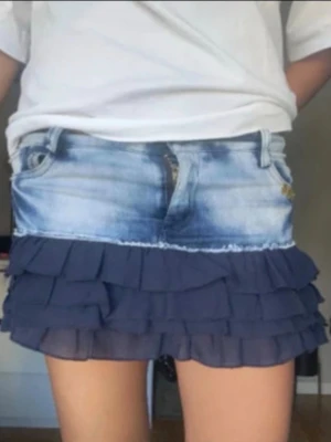 Jeanskjol med volang i blått - Kort kjol med ljusblå jeans upptill och flera lager mörkblå volanger nedtill. Kjolen har klassiska jeansdetaljer som fickor och bälteshällor, samt dragkedja framtill. Perfekt för en trendig och ungdomlig stil.