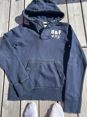 Mörkblå hoodie med tryck och knapp - Mörkblå hoodie med huva och knappdetalj vid halsen. Tryck med texten 'a&f of N.Y.' på bröstet. Stor känguruficka framtill och ribbade muddar vid ärmar och nederkant. Skön och avslappnad stil, perfekt till jeans eller mjukisbyxor. Personen på bilden 165-175 . 50-60kg