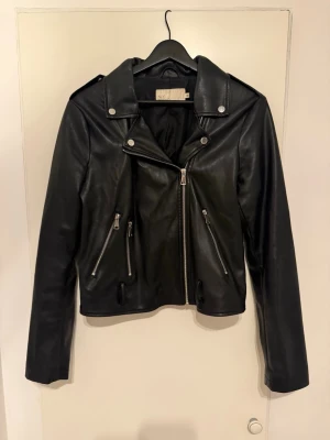 Svart biker skinnjacka med dragkedjor - Snygg svart skinnjacka i klassisk bikerstil med silvriga dragkedjor och axelklaffar. Jackan har snedställd dragkedja framtill, två fickor med dragkedja och detaljerade sömmar. Perfekt för en cool och trendig look. Jackan saknar skärpet i midjan.