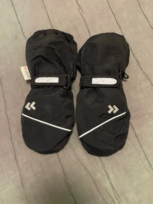 Svarta tumvantar med reflexdetaljer - Säljer ett par svarta tumvantar med reflexdetaljer och justerbart kardborreband vid handleden. Vantarna har förstärkt greppyta i handflatan och elastisk mudd upptill. Perfekta för kalla dagar och utomhusaktiviteter.