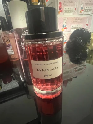 La Fantasía Mango parfym - La Fantasía Mango är en parfym med fruktig doft, levererad i en genomskinlig glasflaska med rosa vätska och svart cylinderformat lock. Flaskan har en modern design och etikett framtill med texten 'Mémoires de la Méditerranée'.