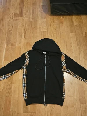 Svart hoodie med rutiga detaljer - Säljer en svart hoodie med dragkedja och huva. Hoodien har klassiska beige, vita och svarta rutiga detaljer längs ärmarna och sidorna. Stora fickor framtill och justerbar dragsko i huvan. Perfekt för dig som gillar streetwear och vill sticka ut lite extra. ÄR S MEN ÄR SON XS