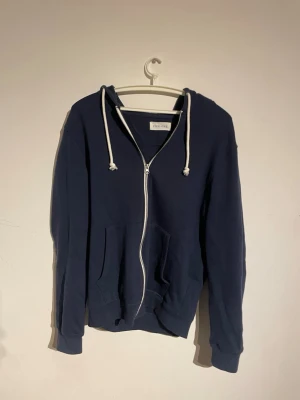 Mörkblå hoodie med dragkedja - Mörkblå hoodie med vit dragkedja och vita snören i huvan. Klassisk modell med två fickor framtill och ribbade muddar vid ärmar och nederkant. Perfekt för en avslappnad stil och enkel att matcha med jeans eller joggers. Nypris 800kr