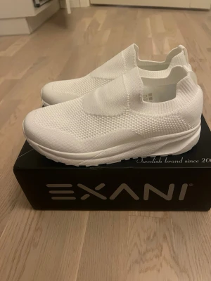 Vita slip-on sneakers från Exani - Stilrena vita slip-on sneakers från Exani med stickad ovandel i mesh för extra andningsförmåga. Skorna har en chunky sula och elastisk mudd runt ankeln för enkel påtagning. Perfekta för dig som gillar minimalistisk och sportig stil.