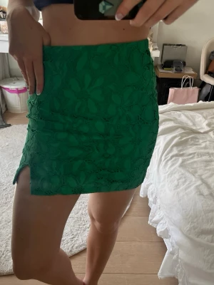 Grön spetskjol från H&M - Säljer en grön kortkjol från H&M med blommigt spetsmönster och liten slits på sidan. Kjolen har en tight passform och är perfekt för dig som vill sticka ut med färg. Supersnygg till både sneakers och klackar!